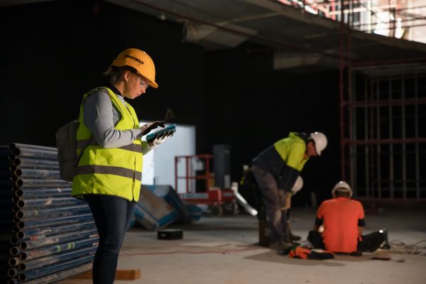 Eine Frau mit Schutzkleidung blickt auf einer Baustelle auf ihr Smartphone, zwei Männer befinden sich im Hintergrund.