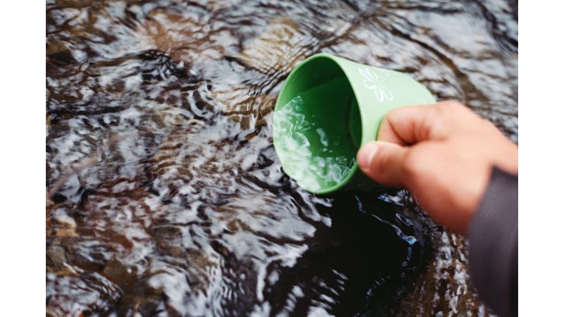 Das Bild zeigt eine Hand, die eine Tasse in ein Gewässer hält.