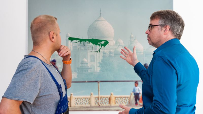 Zwei Männer stehen vor einem Bild des Taj Mahal, auf dem sich ein grüner Farbklecks befindet.