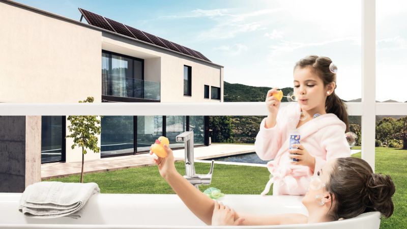 Eine Frau liegt in einer Badewanne, daneben steht ein Mädchen, das Seifenblasen macht. Im Hintergrund steht ein Einfamilienhaus.