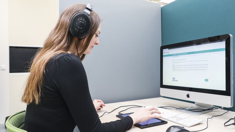 Eine Frau sitzt mit Kopfhörern vor einem Computer an einem Schreibtisch.