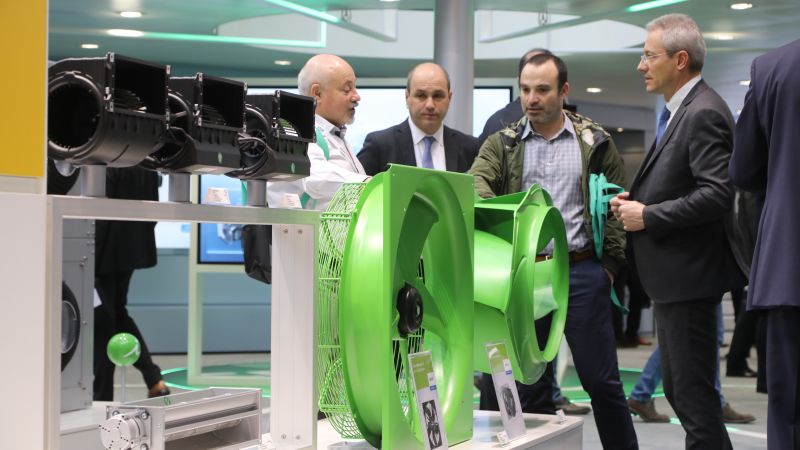 Das Bild zeigt einen Messestand mit raumlufttechnischen Produkten, darunter zwei grüne Ventilatoren, und einige Personen, die diese betrachten.