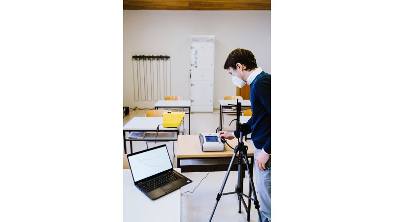 Das Bild zeigt einen Mann mit Atemschutzmaske, der in einem Klassenzimmer mit Hilfe von Messgeräten und einem Laptop eine Messung durchführt. An einer Wand im Hintergrund steht ein Luftreiniger.