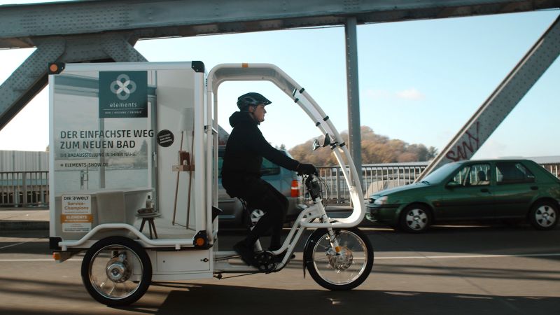 Das Bild zeigt einen Stadtlieferanten auf einem Lastenfahrrad unterwegs auf einer Straße.