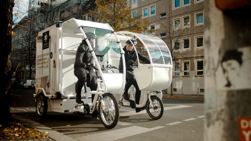 Das Bild zeigt zwei nebeneinander stehende Lastenfahrräder auf einer innerstädtischen Straße. Die Fahrer sitzen auf den Rädern und unterhalten sich miteinander.