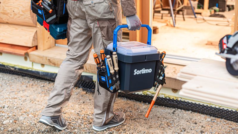 Ein Handwerker läuft und hält dabei eine Box, an der eine Werkzeugtasche hängt, in der Hand.