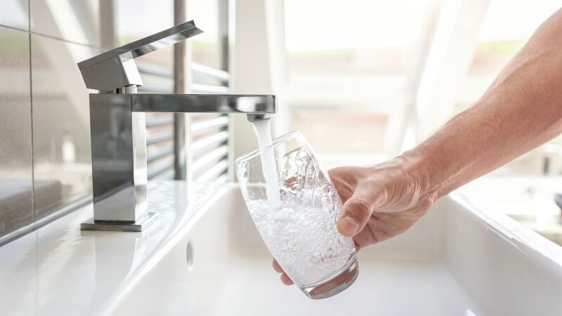 Das Bild zeigt eine Hand mit einem Wasserglas unter dem Wasserstrahl einer Armatur.