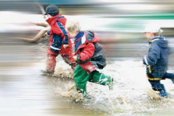 Das Bild zeigt drei Kinder, die in regenkleidung durch eine überflutete Straße laufen.