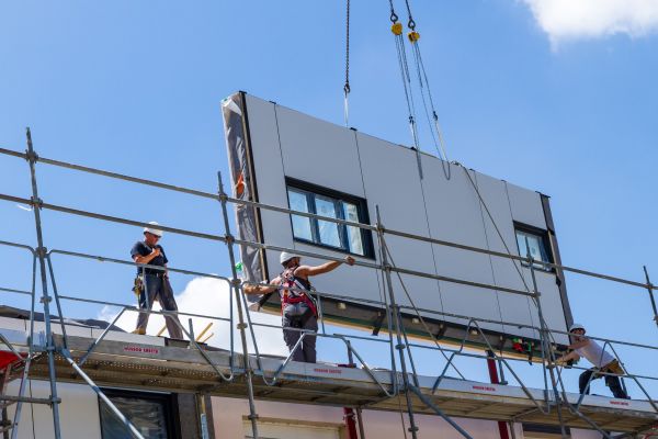 Ein vorgefertigtes Fassadenelement wird auf eine Baustelle gehievt, drei Arbeiter helfen.