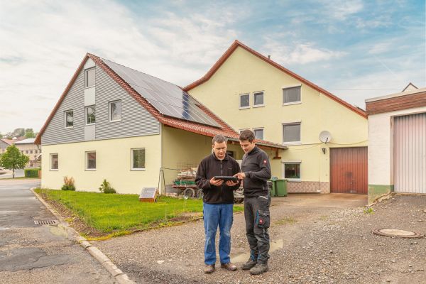 Zwei Männer stehen vor einem Haus und schauen auf ein Tablet.