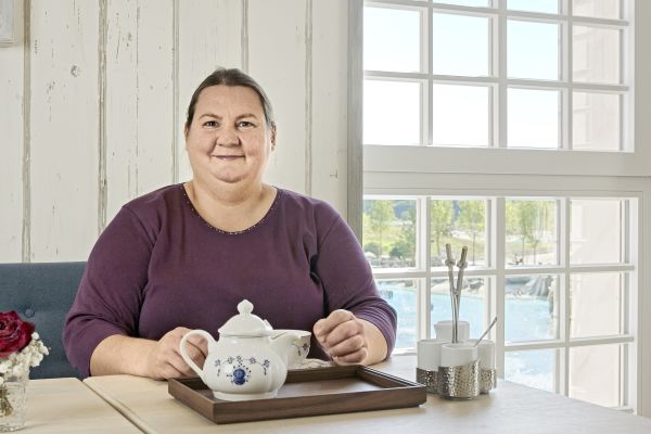 Eine Frau sitzt an einem Tisch, vor ihr eine Teekanne und Tasse.